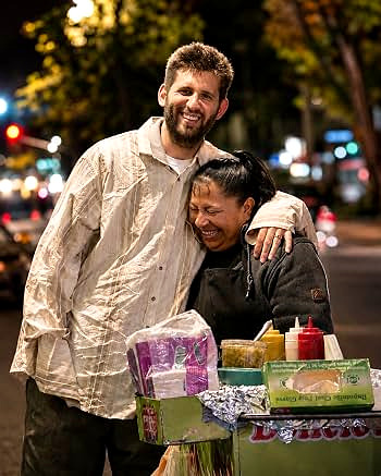 Josh Liljenquist embracing a street vendor at night beside a food cart
